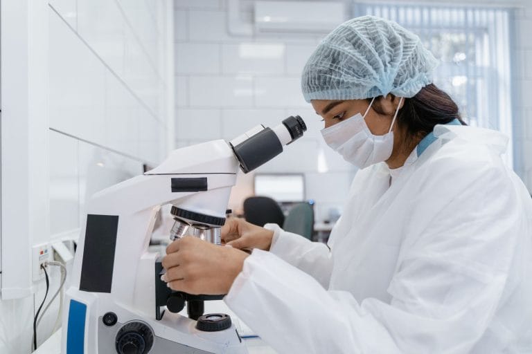 Anvisa seleciona até oito projetos de novos medicamentos para aconselhamento regulatório Cientista usando equipamento de proteção analisa amostras ao microscópio em um laboratório moderno.