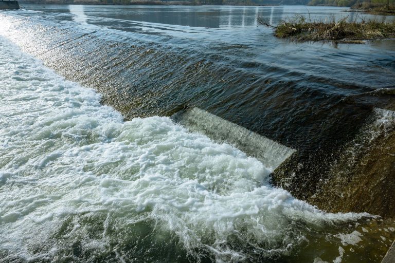 Água escoa por uma barragem em direção a um rio, em uma paisagem natural.
