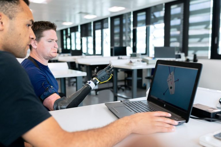 Dois pesquisadores analisam o projeto de uma prótese em um laptop dentro de um laboratório.